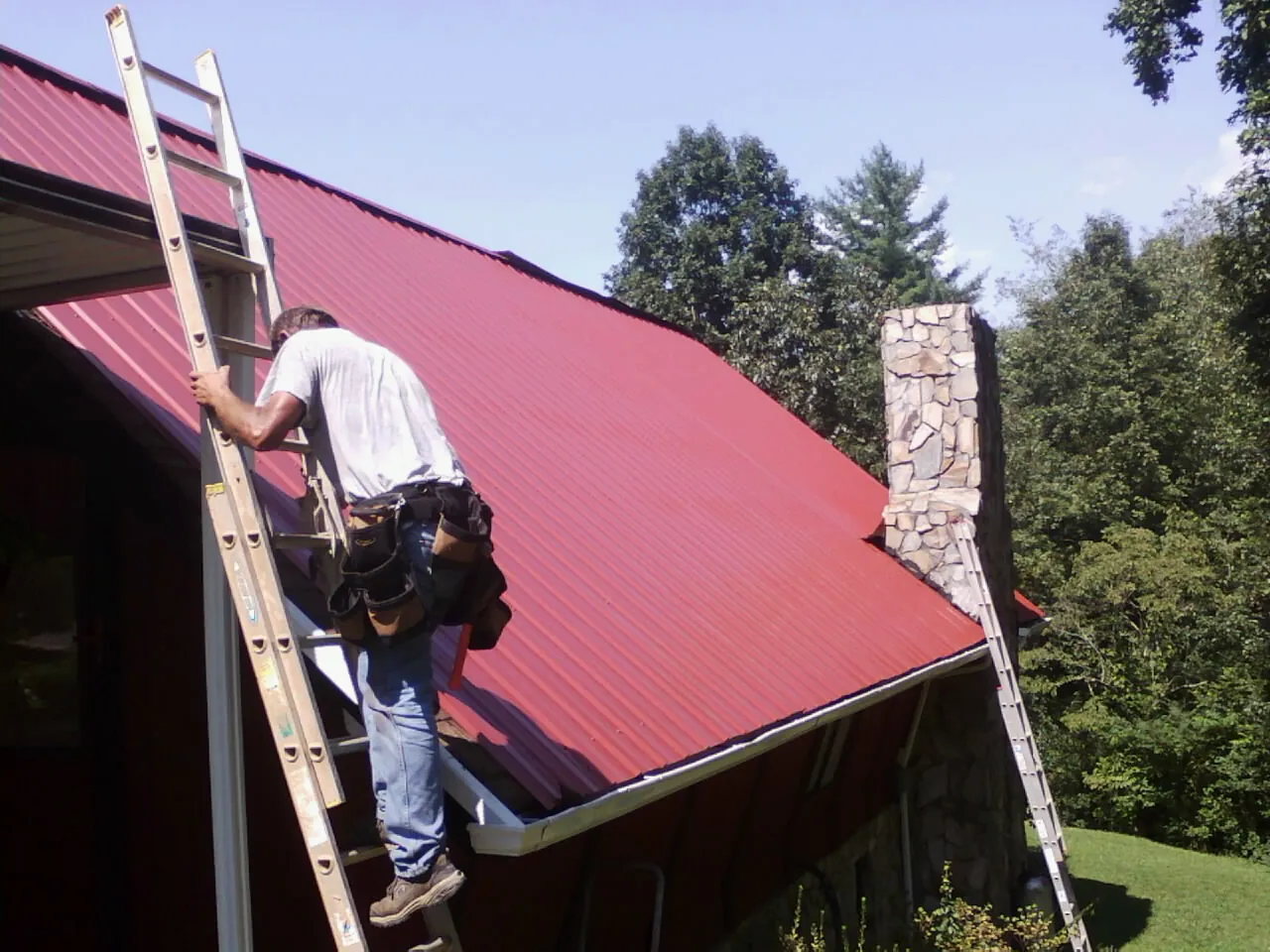 Roofer on a ladder beside a red metal roof and stone chimney — Soffit & Fascia Repair by Roofing Brothers in Payette